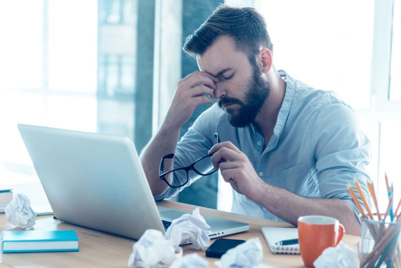 Homme barbu assis à son bureau, visiblement stressé ou fatigué, retirant ses lunettes et se frottant les yeux devant son ordinateur portable, entouré de feuilles froissées. Homme barbu assis à son bureau, visiblement stressé ou fatigué, retirant ses lunettes et se frottant les yeux devant son ordinateur portable, entouré de feuilles froissées.