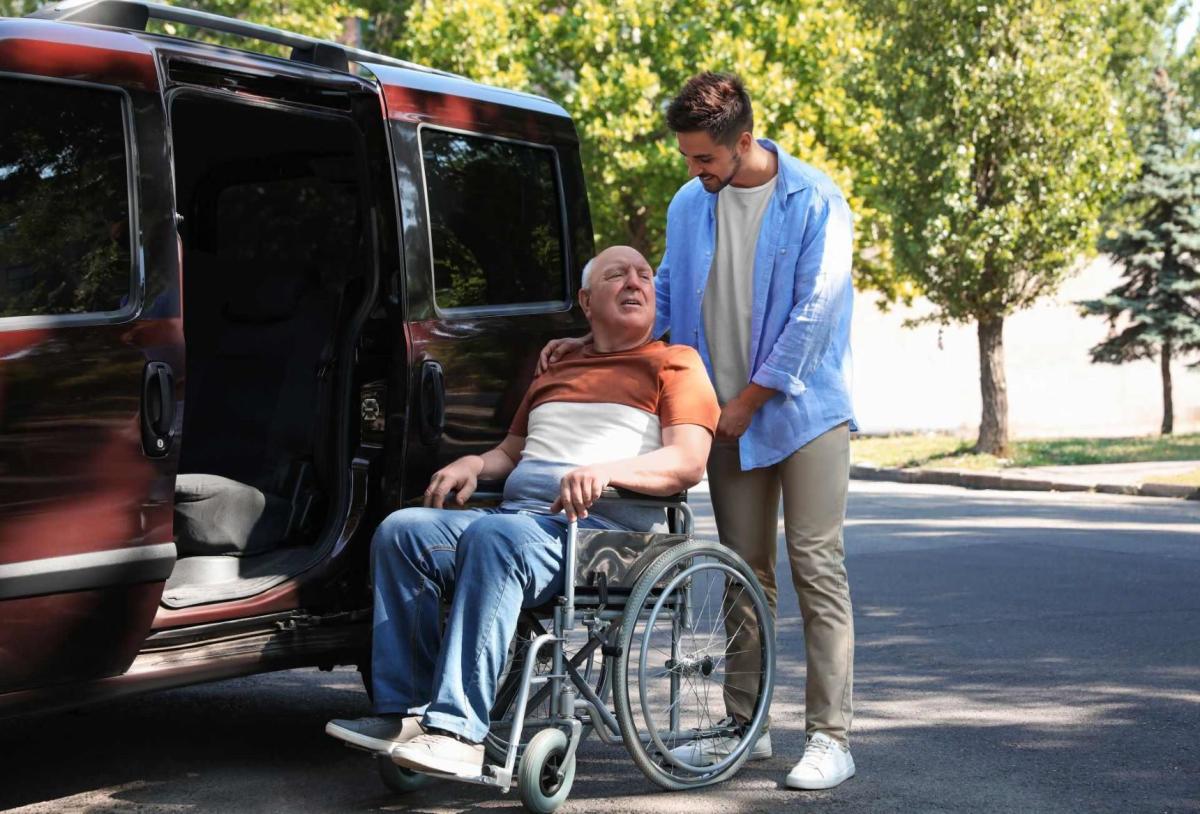 Jeune homme aidant un patient en fauteuil roulant à monter dans une camionnette à l'extérieur /  Jonge man helpt een rolstoelpatiënt in een openluchtbusje