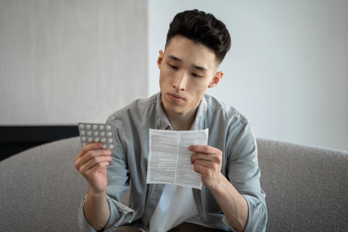 Jeune homme tenant une plaquette de médicaments pour soulager la douleur assis sur un canapé à la maison /Jongeman die een blisterverpakking met medicijnen vasthoudt voor pijnverlichting terwijl hij thuis op een bank zit