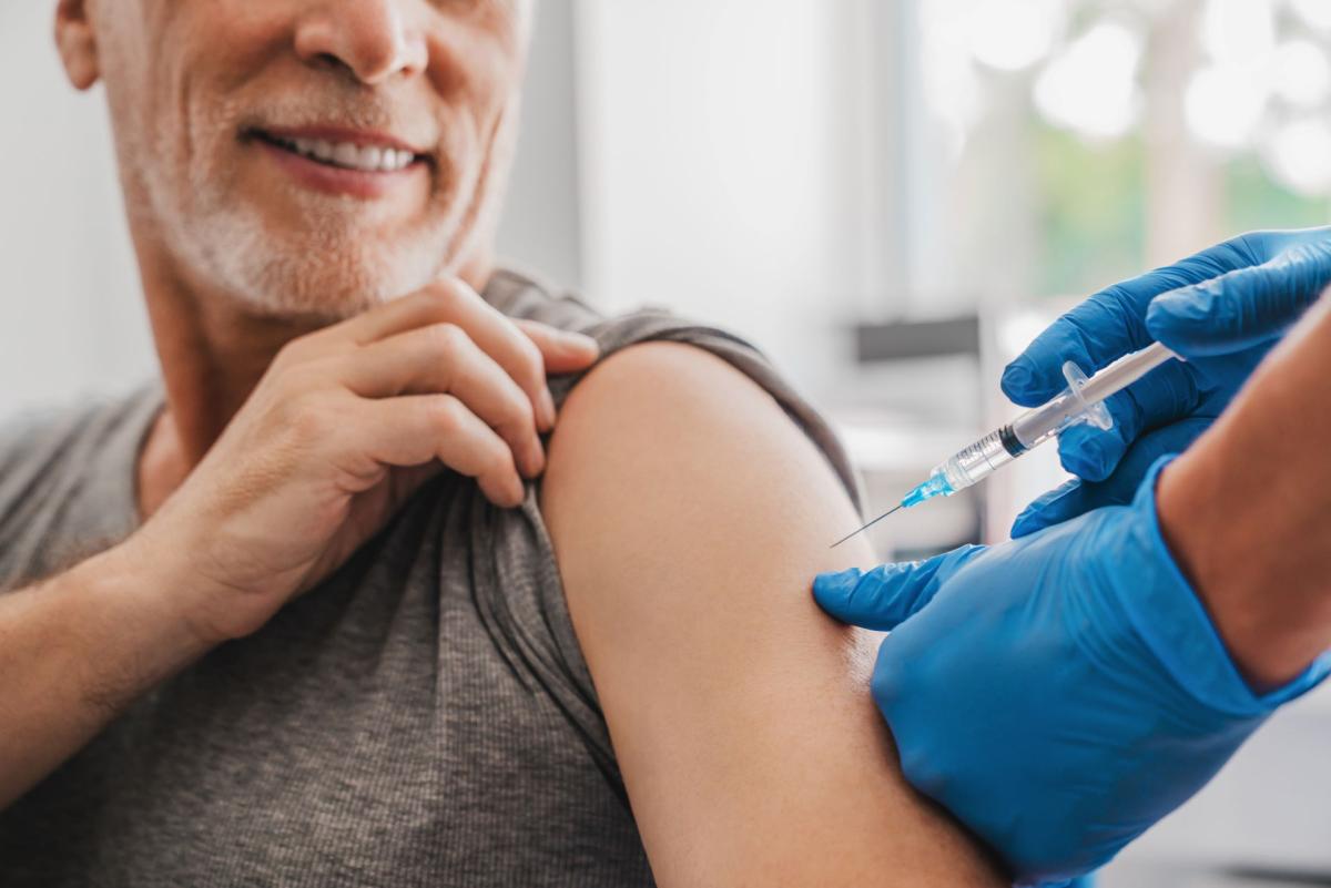 Homme souriant recevant une injection dans le bras par un professionnel de santé portant des gants bleus, illustrant un acte de vaccination Homme souriant recevant une injection dans le bras par un professionnel de santé portant des gants bleus, illustrant un acte de vaccination