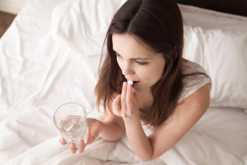 Jeune femme assise dans un lit et prenant un comprimé avec un verre d'eau / Jonge vrouw zittend in bed neemt een tablet met een glas water