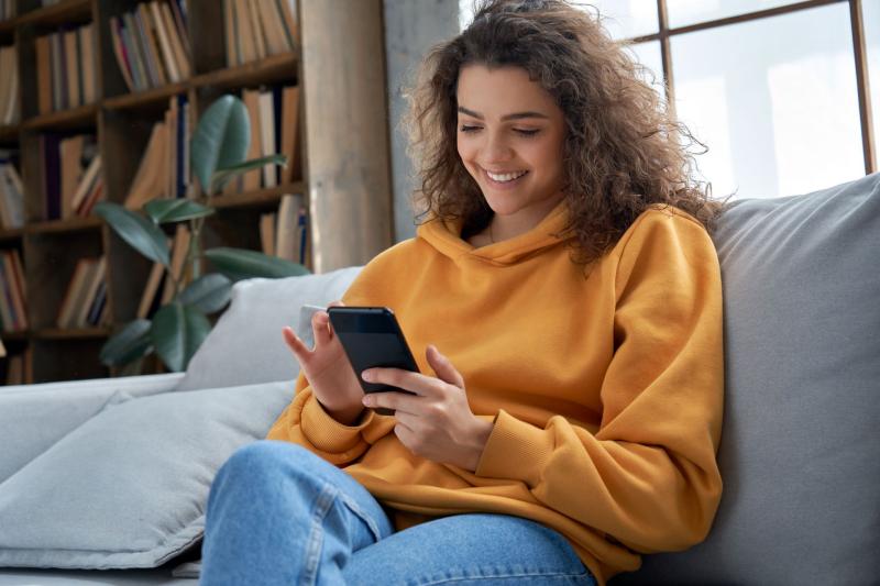 Fille souriante assise dans son divan avec son téléphone en main/Lachende meisje zittend op haar bank met haar telefoon in de hand/