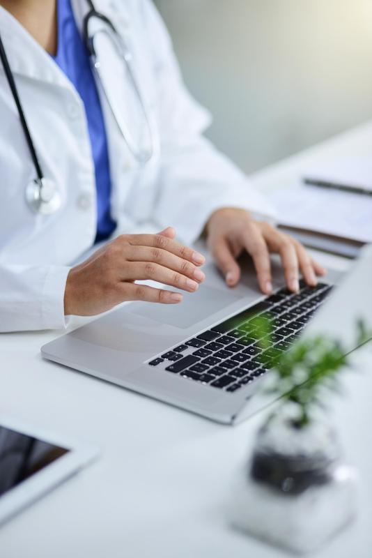 Un médecin en blouse blanche, équipé d’un stéthoscope, utilise un ordinateur portable dans un environnement de bureau médical.