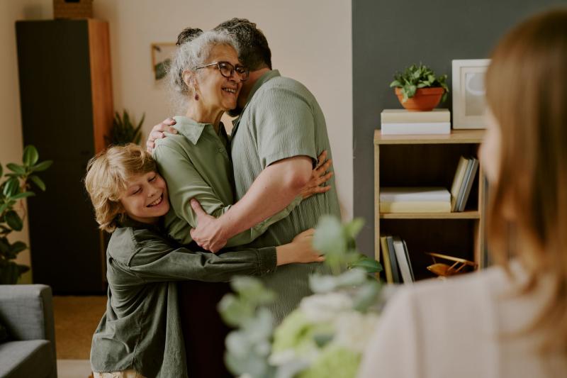 Moment d’affection intergénérationnelle : une femme âgée souriante enlace un homme adulte et un enfant dans un cadre familial chaleureux, sous le regard attendri d'une autre personne Moment d’affection intergénérationnelle : une femme âgée souriante enlace un homme adulte et un enfant dans un cadre familial chaleureux, sous le regard attendri d'une autre personne